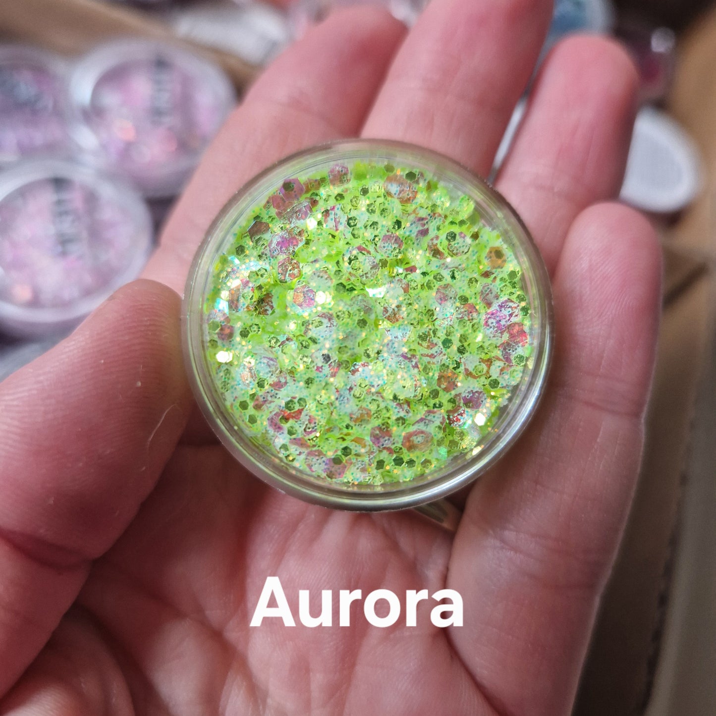 Aurora Glitter Cream i neon gul-grøn med rosa og sølv glitter fra Fusion Body Art, holdt i hånd