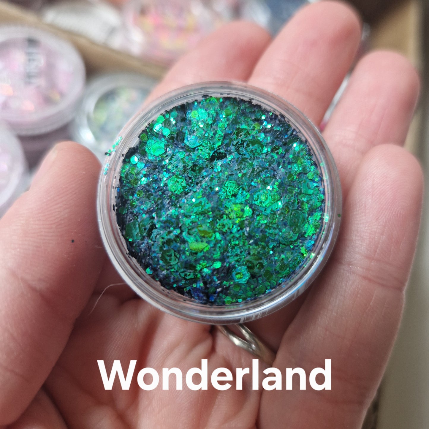 Wonderland Glitter Cream i grøn og blå nuance holdt i hånd, viser skinnende glittereffekt